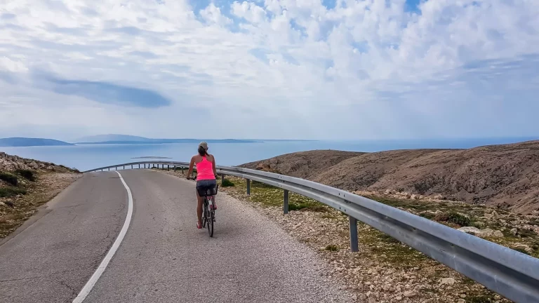 Ein Mädchen in einem sportlichen Outfit, fahren eine steile Straße hinunter auf einem Fahrrad. in der unteren Mittelmeer. Das Mädchen genießt die Fahrt. Dicke Wolken darüber. Aktiver Urlaub. Schöne Küstenlinie von Kroatien